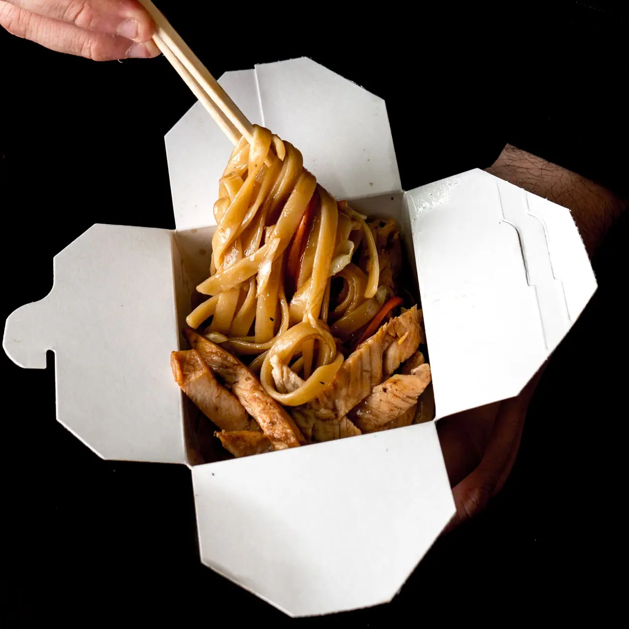 Noodles in a white takeout box lifted with chopsticks